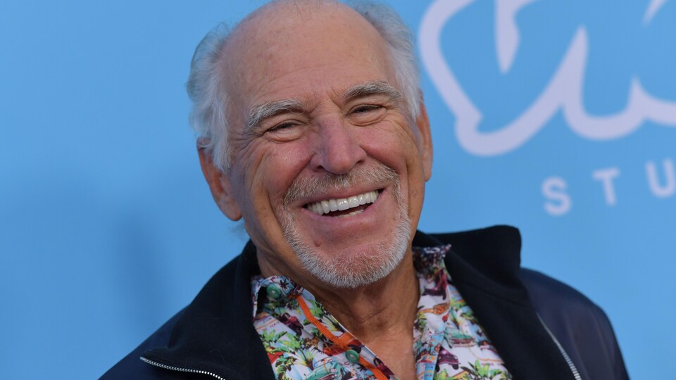 Le chanteur américain Jimmy Buffett, en chemise hawaïenne et veste de sport, sourit à la caméra devant une toile bleue ciel.