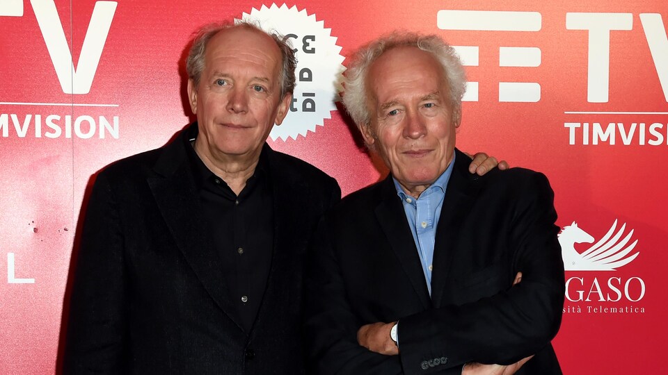 Les frères Jean-Pierre et Luc Dardenne debout devant un mur rouge.