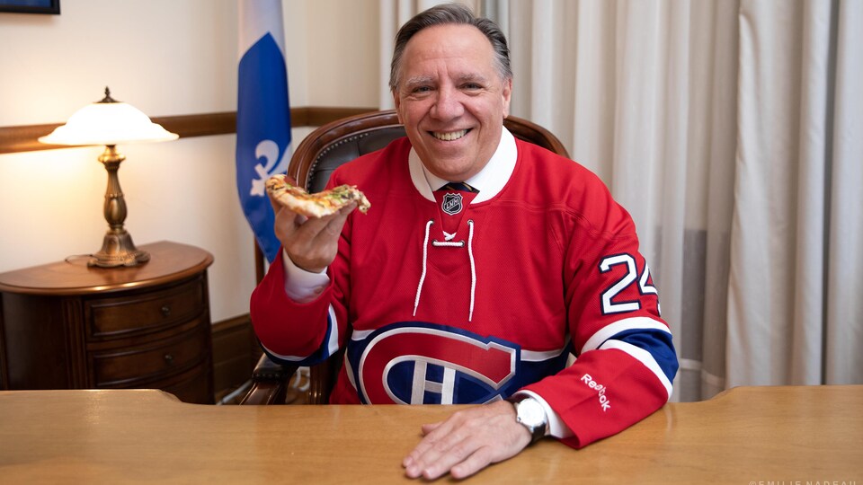 François Legault porte un chandail du Canadien et tient une pointe de pizza.