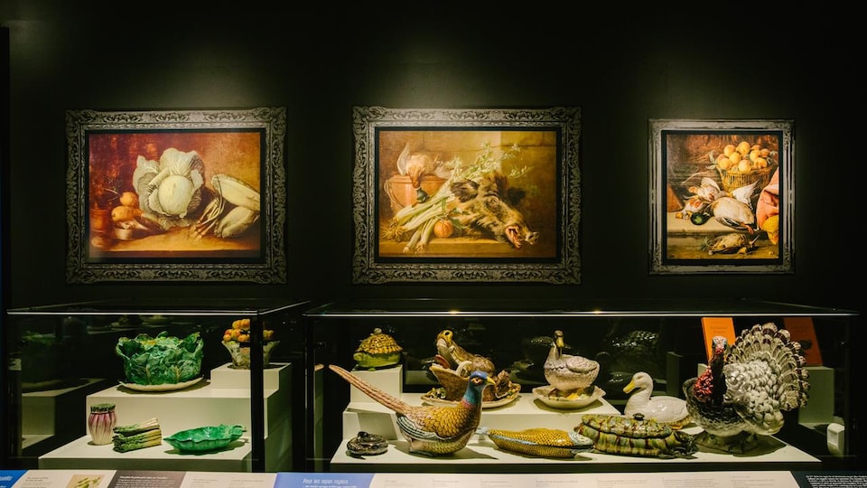 Des plats de service de fantaisie sur un présentoir couvert de verre, surplombé de trois peintures représentant des plats français typiques.