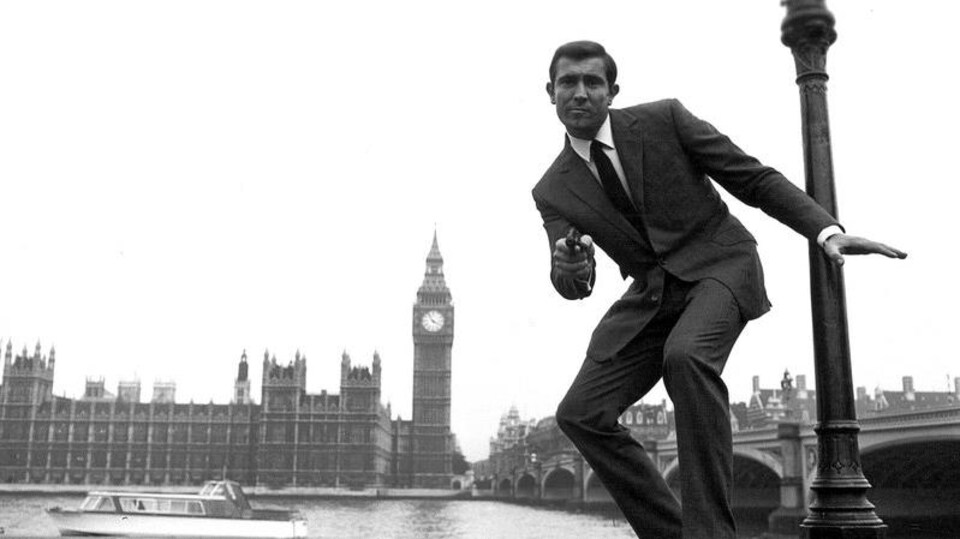 L'acteur se tient debout au bord de la Tamise avec la tour de Big Ben en arrière-plan et il s'apprête à tirer avec un revolver.