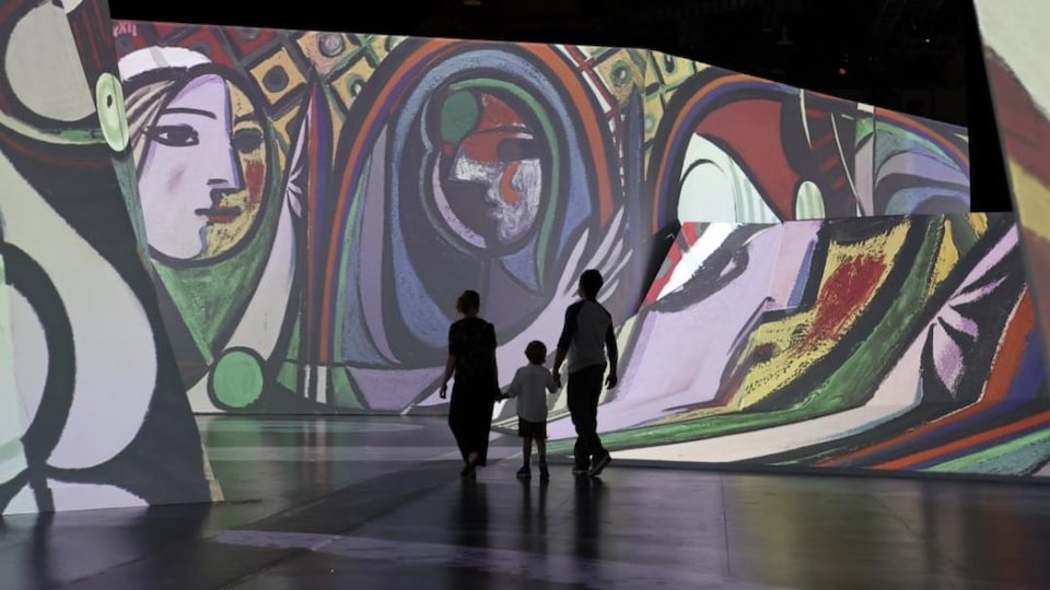 Un homme, une femme et un enfant se promènent dans l'exposition, entourés d'œuvres de Picasso projetées sur les murs.