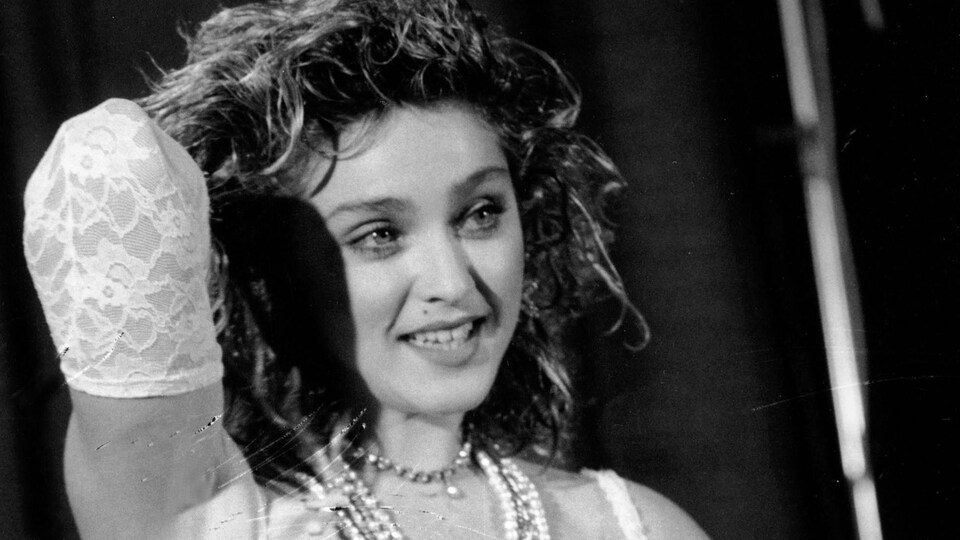 La chanteuse pose le bras droit levé en souriant aux photographe. Elle porte des chapelets, une robe de mariée et une ceinture avec une boucle Boy Toy.