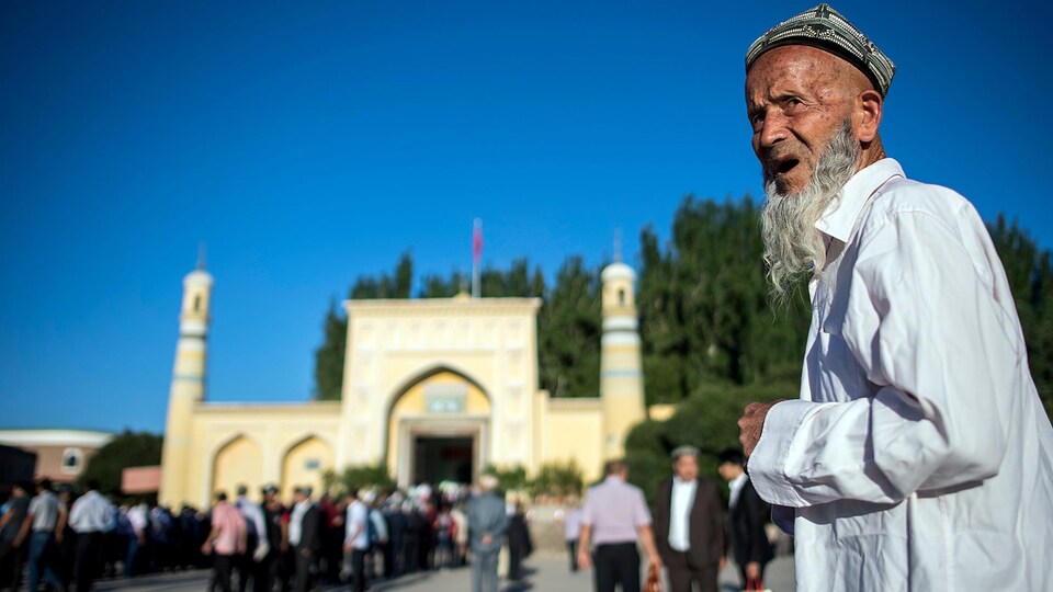 À l'avant-plan, un homme est debout devant une mosquée. Plusieurs personnes sont massées à l'entrée du bâtiment.