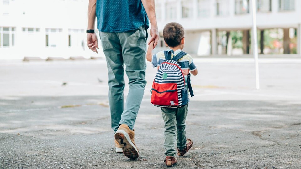 Un homme tient un jeune enfant par la main dans une cour d'école.