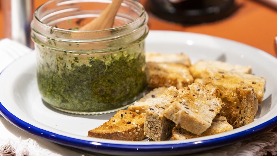 Un petit pot de pesto aux fanes de carottes posé sur une assiette, à côté de croûtons.