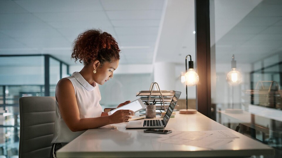 Une femme travaille sur une tablette informatique; un ordinateur est posé sur son bureau.