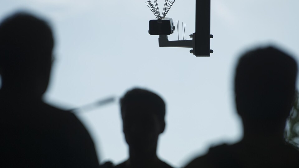 Une caméra est fixée dans les airs alors que des passants, en contrejour, circulent.