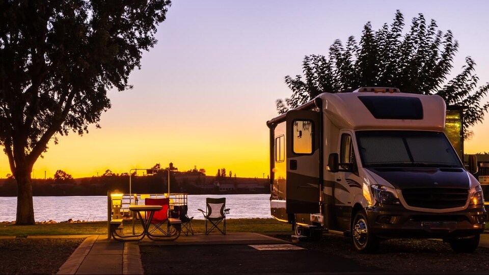 Photo prise au coucher du soleil d'une véhicule récréatif stationné au bord d'un plan d'eau avec des chaises et une table de camping sorties.