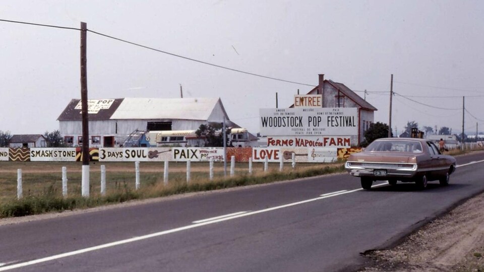 Photo de l'entrée du site, dans un champ au bord d'une route, avec des panneaux sur lesquels sont inscrits « Entrée Woodstock Pop Festival ».