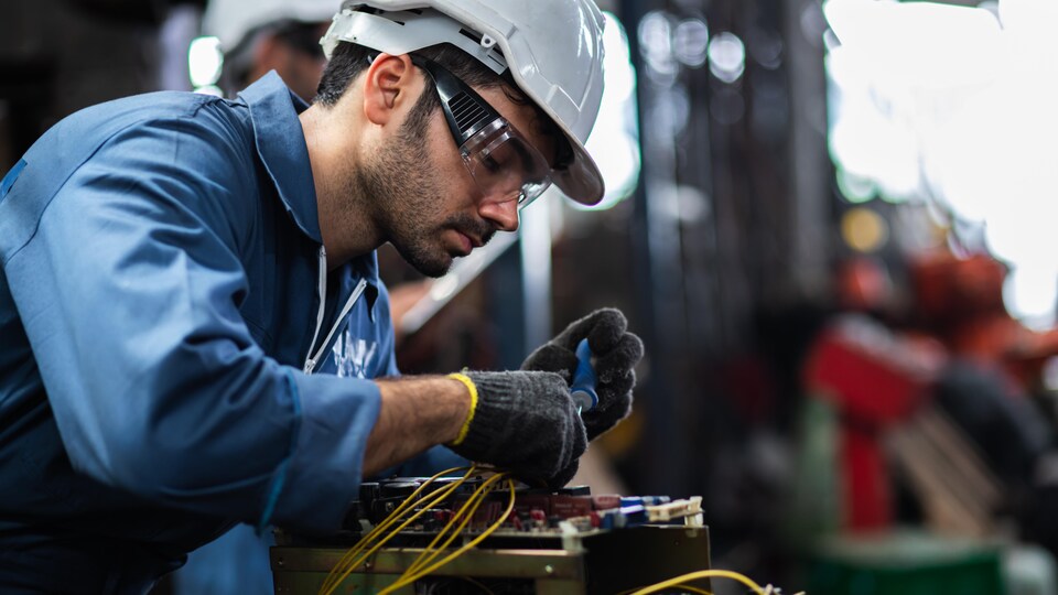 Un travailleur dans une usine, avec un casque, des gants et des lunettes de protection, assemble les composantes d'un appareil. 