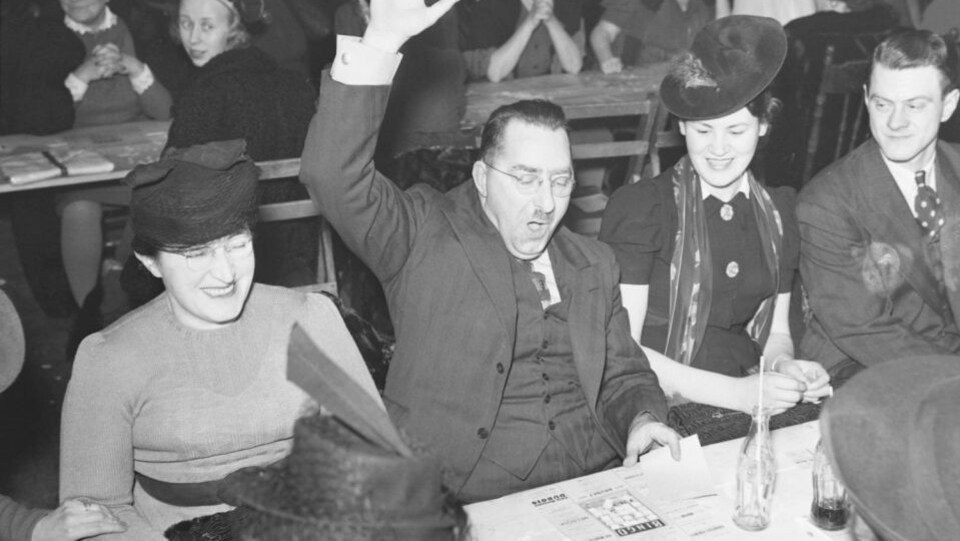 Un gagnant de bingo, assis avec d'autres joueurs, lève la main. Cette partie a lieu au 4555, avenue Notre-Dame-de-Grâce à Montréal en 1941.