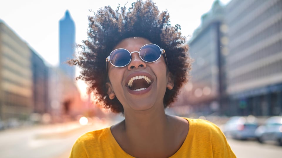 Une jeune femme avec des lunettes soleil sourit à grandes dents.