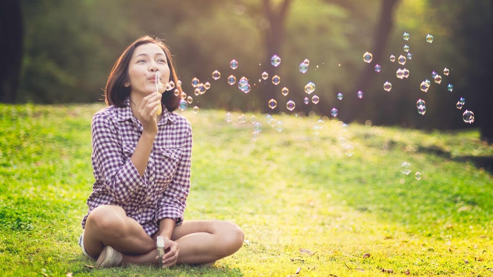 Assise en tailleur, une jeune femme fait des bulles. 