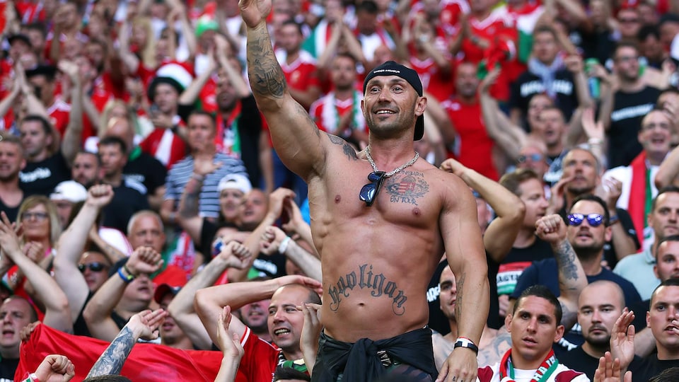 Dans une foule assise, un homme torse nu qui porte un tatouage du mot Hooligan lève le bras gauche.
