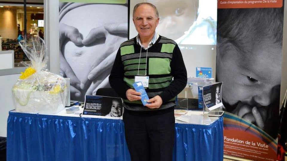 Un homme debout regarde la caméra en souriant devant un kiosque et en tenant une brochure dans ses mains.