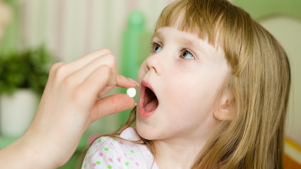 Une main tient une pilule blanche devant la bouche ouverte d'une jeune fille.