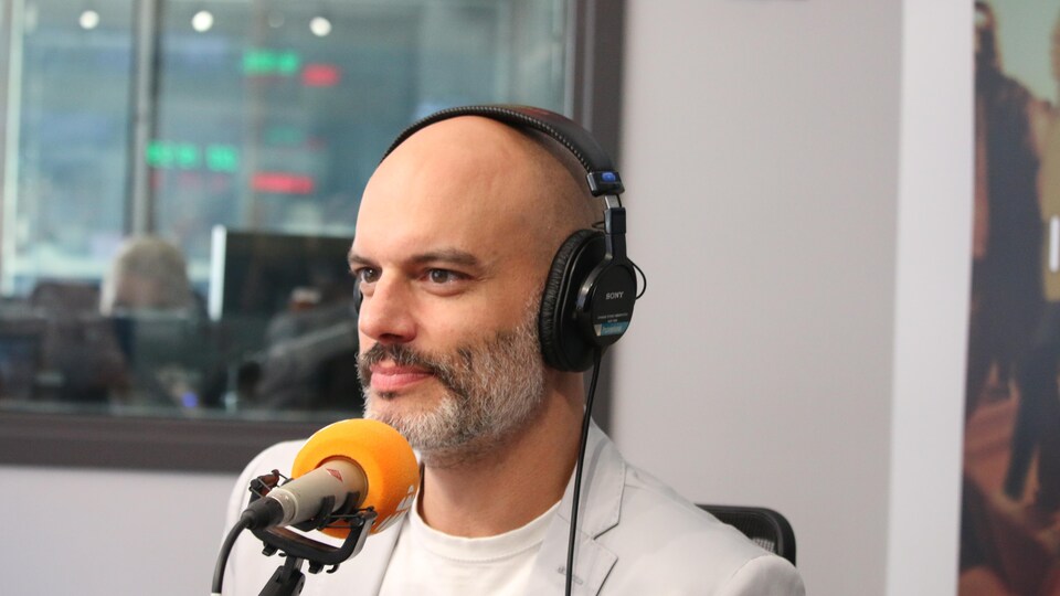 Un homme habillé de couleurs pâles est devant un micro au studio radio.