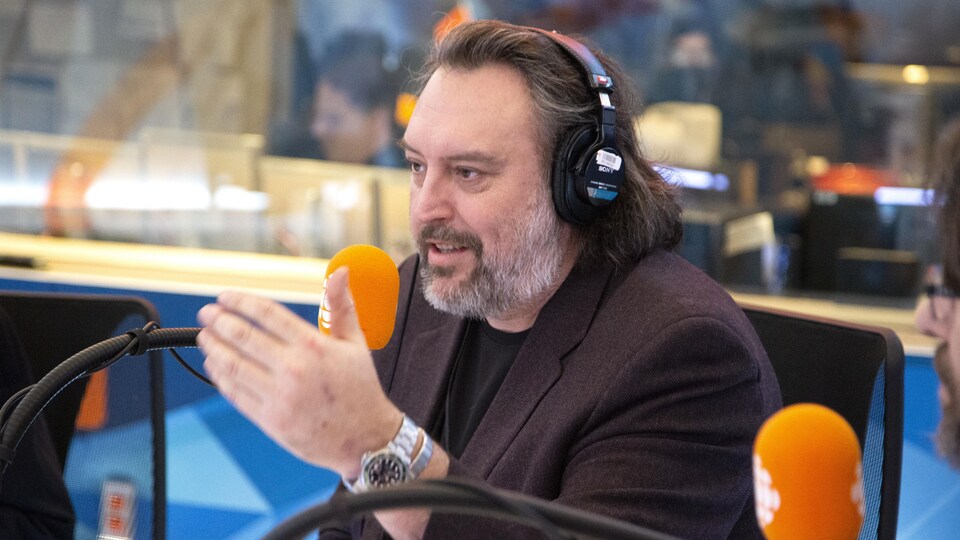 Assis derrière un micro, il lève la main gauche en parlant.
