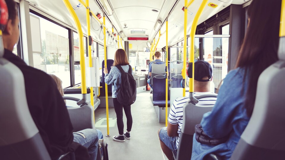Une adolescente est debout dans l'allée pendant un trajet en bus. D'autres adultes sont assis de dos, le regard vers l'avant du véhicule.