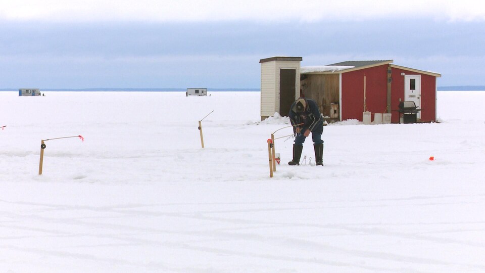 Canne à pêche à la main, un homme est penché au-dessus d'un trou dans la glace devant sa cabane.