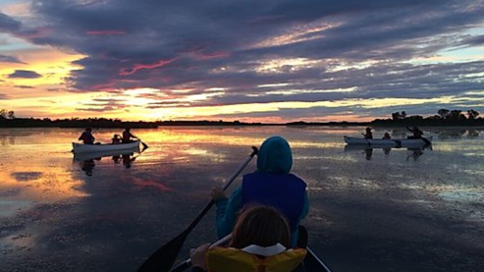 Des enfants et des adultes dans trois canots sur un lac, au soleil couchant