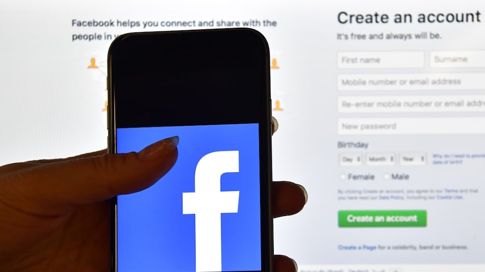 Une main tient un téléphone intelligent montrant le logo de Facebook devant l'écran d'un ordinateur qui affiche la page d'accueil de Facebook.