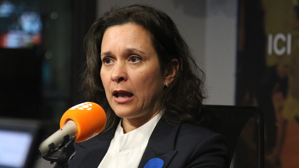 Une femme aux cheveux bruns en tailleur noir et chemise blanche parle au micro.