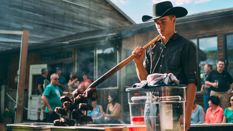un jeune homme avec un chapeau de cowboy fait griller des steaks au barbecue avec une fourche