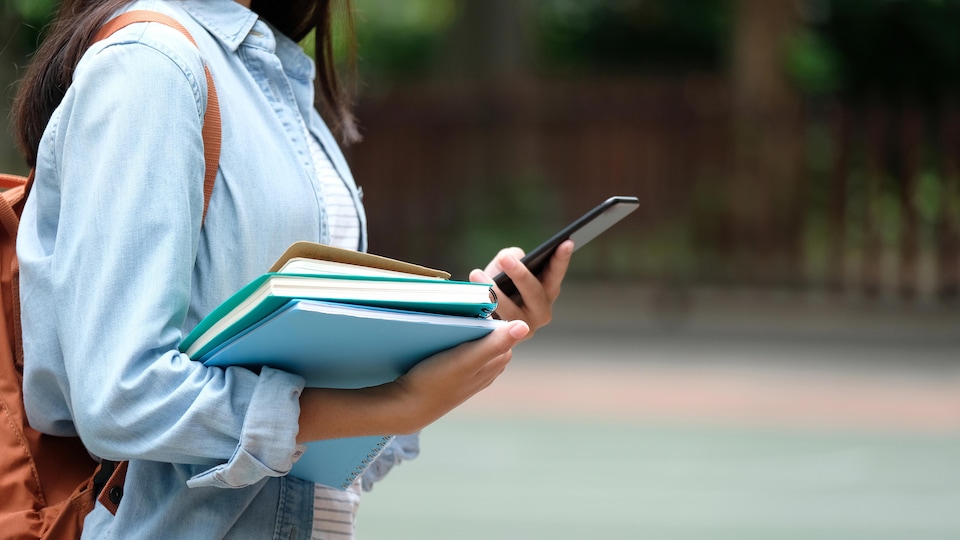 Une femme regarde son cellulaire en tenant ses livres d'école.