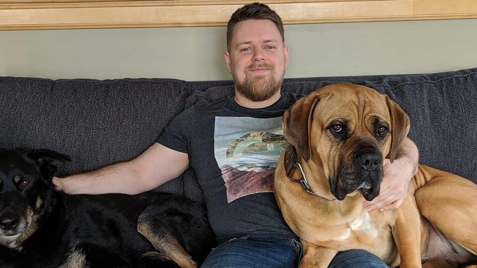 Un homme est assis sur un sofa avec deux chiens d'assez grande taille couchés sur le sofa près de lui.