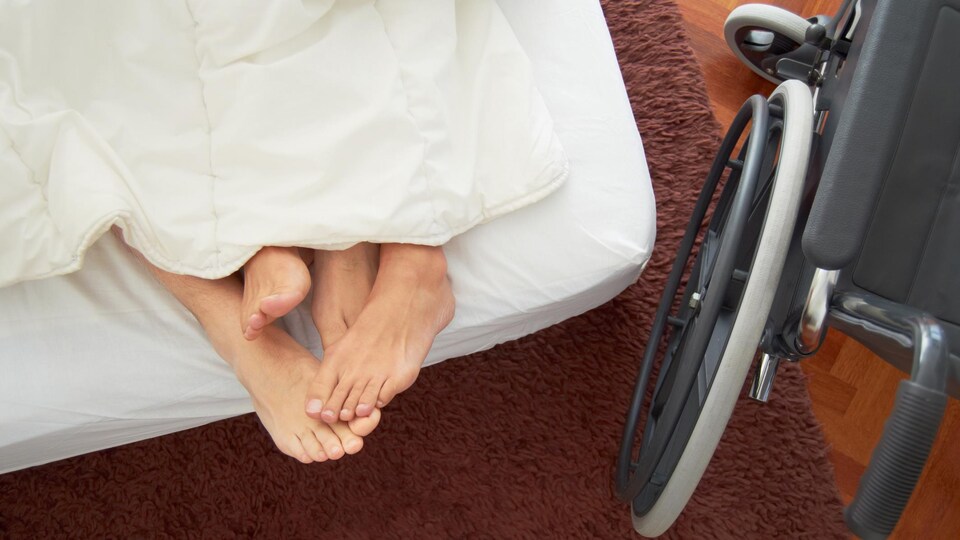 On voit quatre pieds emmêlés dans un lit et une chaise roulante à côté.