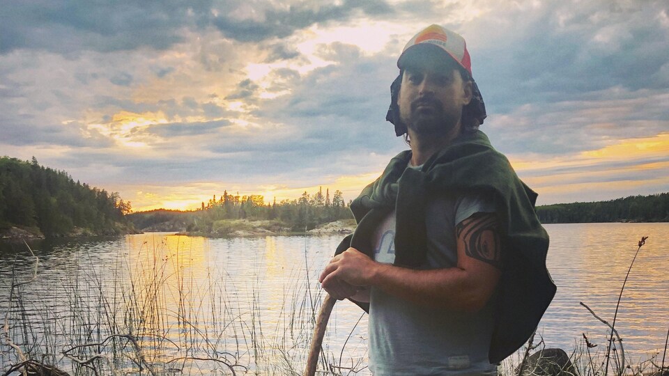 Chris Gaudry, réalisateur métis, musicien et passionné de nature est debout sur les rives d'un étendu d'eau.