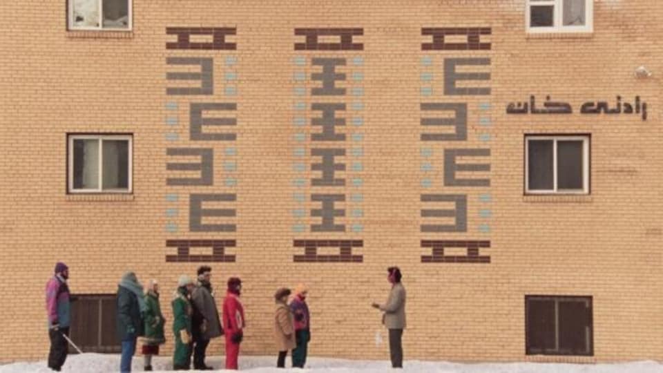 Une image extraite du film. Neuf personnages debouts devant un bâtiment décoré de symboles et de mots. 