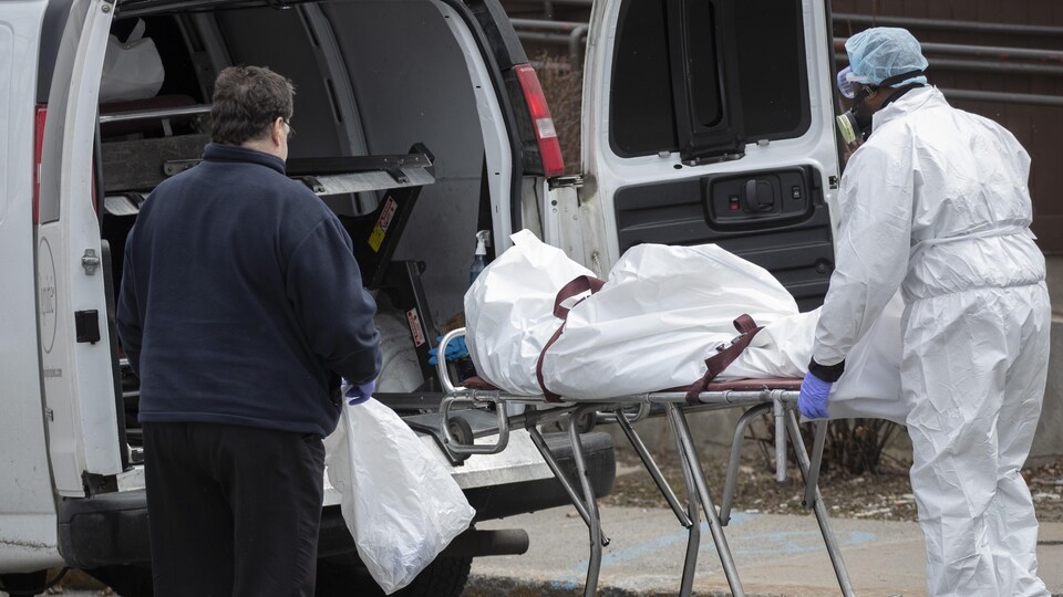 Un préposé masqué et en combinaison protectrice transporte un cadavre sur une civière.