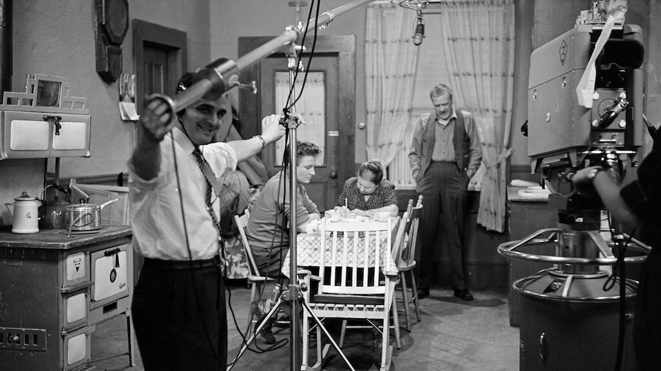 Vue d'ensemble du plateau de tournage avec le preneur de son et la caméra filmant une scène dans la cuisine des Plouffe avec les comédiens Pierre Valcourt, Amanda Alarie et Paul Guèvremont.