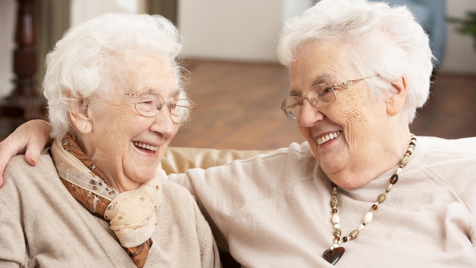 Deux femmes âgées souriant en se regardant. L'une enlace l'autre autour des épaules.