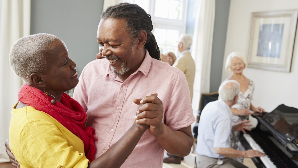 Un couple danse à l'avant-plan pendant qu'un autre est à l'arrière autour d'un piano.