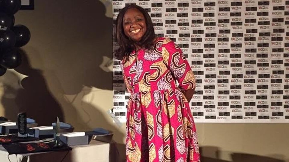 Une femme souriante porte une longue robe rose et jaune faite de tissu au motif africain.