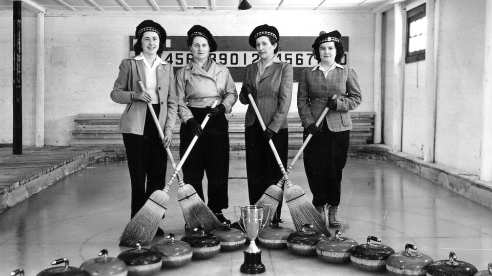 Quatre femmes se trouvent sur une glace destinée au curling. Elles tiennent chacune un balai et posent devant les pierres et une coupe qu'elles ont remportée lors d'un championnat provincial en 1949.