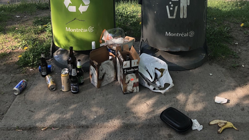 Des bouteilles de bière, un contenant en plastique, une peau de banane et deux caisses de bière au pied d'une poubelle de la Ville de Montréal.