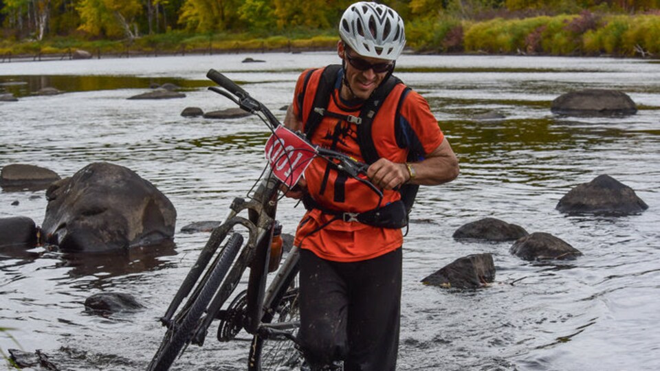 L'athlète Samuel Daigle porte son vélo alors qu'il traverse un cours d'eau.