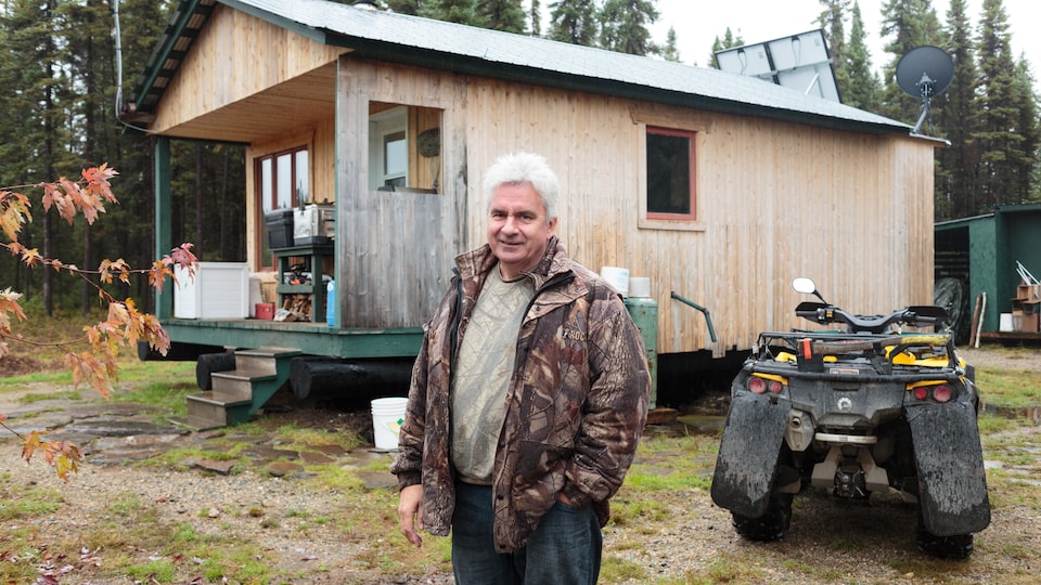 Denys Tanguay pose devant sa résidence. Elle est en bois, deux panneaux solaires trônes sur la toiture, un véhicule tout terrain est à côté de la maison.
