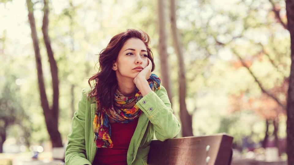 Une femme, l'air déprimé, est assise sur un banc de parc, un jour d'été. 