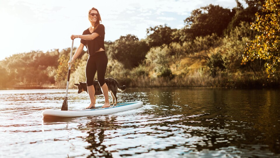 Une belle jeune femme adulte profite d'un moment paisible sur l'eau avec son paddle board et son chien de compagnie. Le soleil illumine la scène, projetant une lueur dorée.
