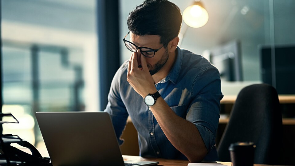 Un homme se tient le nez en signe d'épuisement devant son ordinateur portable posé sur une table.