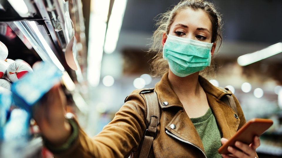 Une femme portant un masque prend un produit dans sa main droite pendant qu'elle se trouve dans un commerce.
