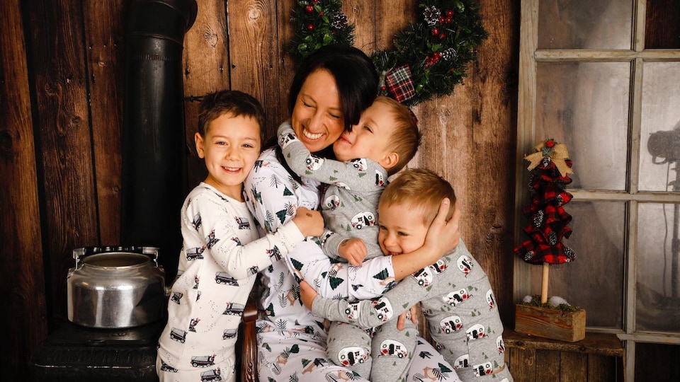 Une mère est assise près d'un poêle à bois. Un de ses enfants est sur ses genoux, deux autres sont de part et d'autre d'elle et la serrent dans leurs bras. Ils sont tous en pyjama. 