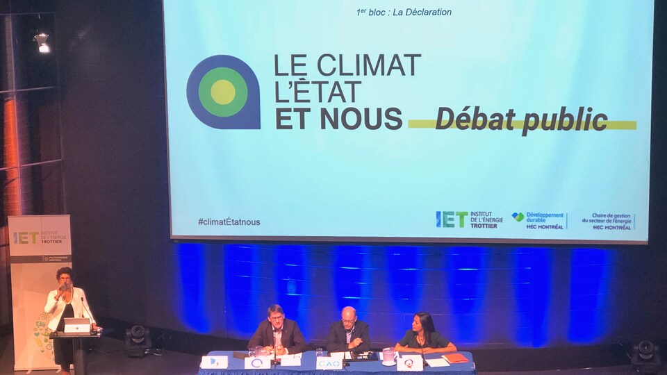 Trois représentants de partis politique différents assis à une table devant public, couronnés d'un écran géant où il est inscrit « Le climat, l'état et nous », Débat public.