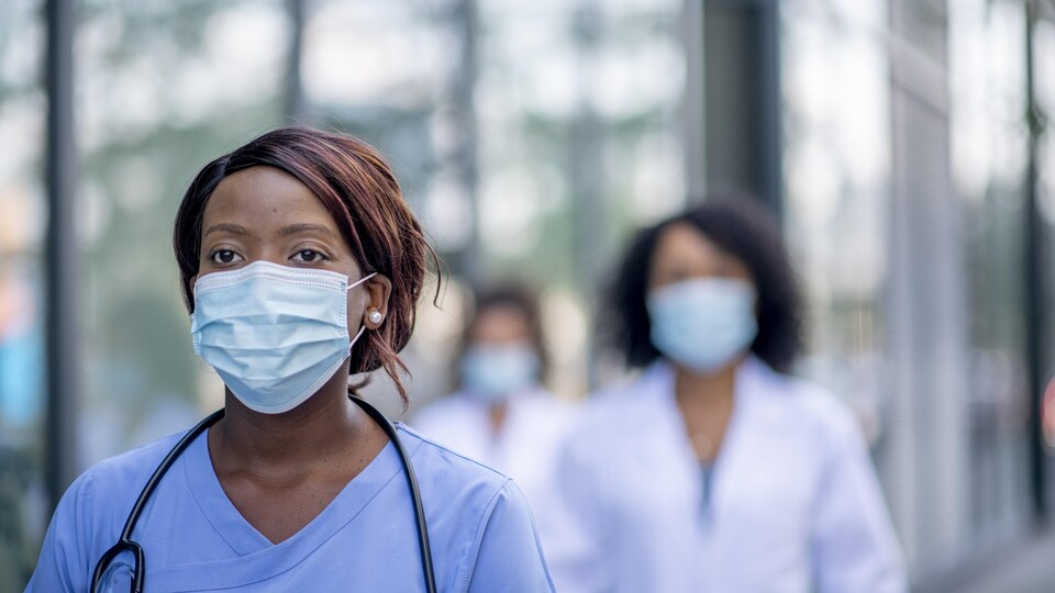 Une infirmière masquée marche à l'extérieur le long d'un établissement de santé, suivi par deux collègues.